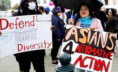 A Black person holding a sign that reads defend diversity standing next to an Asian person holding a sign that reads Asians 4 affirmative action.