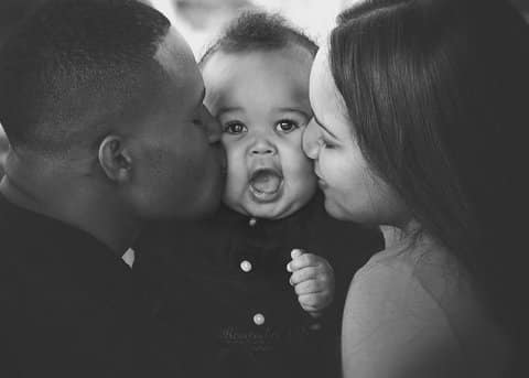 Black-and-white photo of a biracial baby looking at the camera. A Black man and a light-skinned woman are on each side of the baby, kissing his cheek.