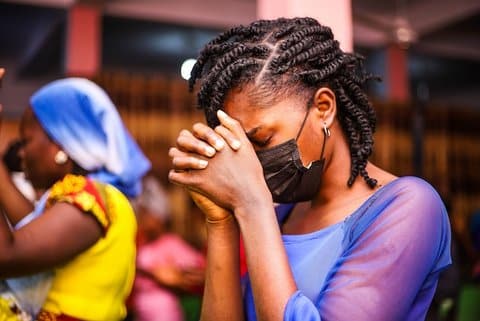 A young Black woman with her eyes closed, hands clasped in front of her face and head bowed in apparent prayer. She is wearing a black medical mask