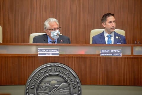 Two Latino men in suits in a government meeting. Placards identify them as Eduardo Martinez, mayor, and Cesar Zepeda, council member district 2. A seal in front of them says Richmond California. One man is wearing a surgical mask with a poop emoji on it.