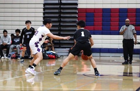 A boys basketball player with the ball and his back to the camera next to an opponent reaching toward him as a referee watches.