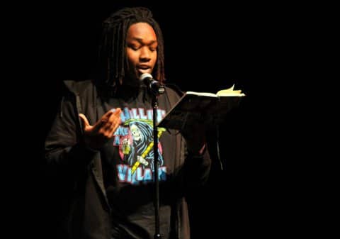 A black boy reading from a book at a microphone. Part of the design on his shirt features a cartoon design of the grim reaper.