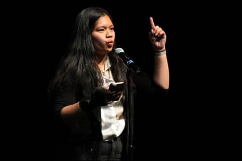 an asian girl at a microphone with a smartphone in one hand and the other hand raised with her index finger pointing upwards. Her lips are pursed, apparently mid-speech
