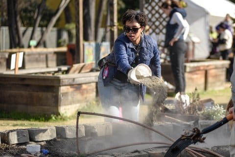 A young black woman wearing sunglasses and hoop earrings throws soil from a bucket while volunteering at a greenery event