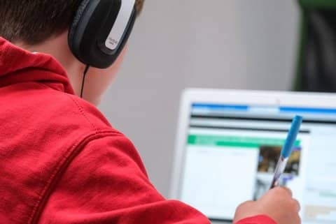 Over the shoulder shot of a boy wearing a red hoodie and headphones looking at a laptop screen, pen in hand. What's on the screen is out of focus