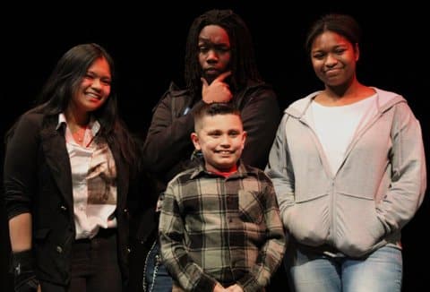 An asian girl, black boy, latino boy and black girl on a stage. The latino boy appears to be in grade school and the others to be teens.