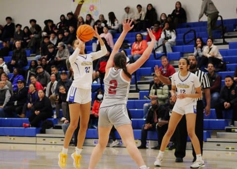 A girls basketball player jumps to shoot the ball over an opponent's upstretched arms with a teammate, referee and fans looking on