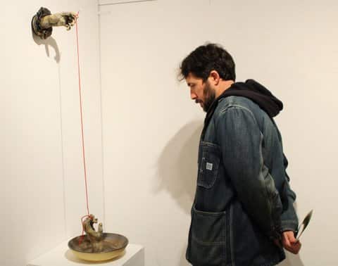 A man looks at an art piece with one reaching up from a shallow bowl and one hand reaching out from the wall. They are connected by a red cord tied to the fingers.