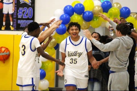 A smiling boys basketball player runs between rows of teammates on either side of him. Many have their arms raised or extended out. There are blue, yellow and gold balloons.