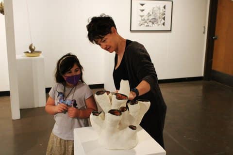 A girl wearing a protective mask and a woman look at a sculpture that is painted white and has tubular branches extending upward. What is visible of the inside is a reddish color and ringed with black. It looks like something you might see in the ocean.