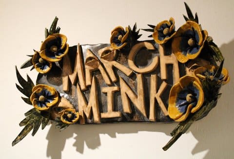 A sculpture of the words "march mink" on a board surrounded by sculpted flowers and feathers. An eye is sculpted into the letters A and R.