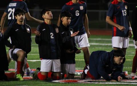 High school soccer players watching and waiting on grass. Some are standing and some on the ground. One is on on knee. Two are on their knees with one with his eyes closed and face and palms turned upward. One is on his hands and knees.