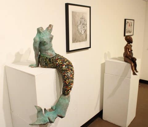 Sculpture of a mermaid that is headless but would have had two heads and a sculpture of a bronze child each sitting on their own display stands with framed artwork on the wall behind them.