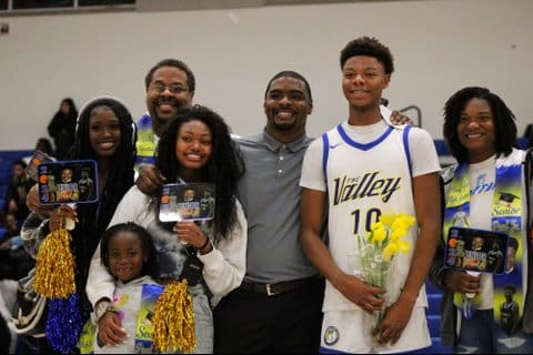 A black teen boy in basketball uniform, smiling, holding a bouquet of yellow flowers and standing with a black woman, three black girls and two black men