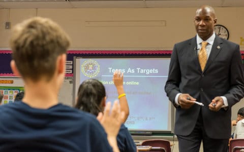 A tall black man in a suit in a classroom looks toward two students seen from behind who are sitting at their desks with their hands raised. A slide projected on the screen says "teens as targets" and has the FBI logo.