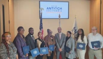 Two black women, a black man, two more black women, another black man, two more black women and a white man standing in a row. Behind them are furled U.S. and Antioch flags and a screen that reads city of Antioch california