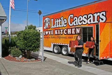 Two people carrying stacks of pizza boxes out of a large truck painted orange that says on the side Little Caesars Love Kitchen serving to make a difference since 19. A man's head is blocking the rest of the year
