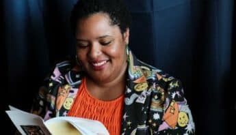 A smiling black woman reading a zine. She is wearing an orange top, green hoop earrings, and a jacket decorated with winking smiley faces, stars, rainbows and other colorful designs.