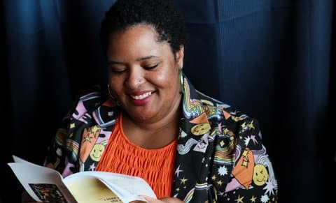 A smiling black woman reading a zine. She is wearing an orange top, green hoop earrings, and a jacket decorated with winking smiley faces, stars, rainbows and other colorful designs.
