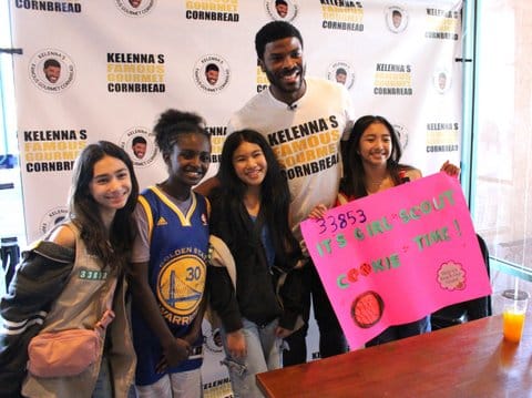 A tall black man wearing a T-shirt that reads Kelenna's famous gourmet cornbread in front of a backdrop that says the same with four girls. One girl is wearing a golden state warriors jersey and one is holding a sign that reads it's girl scout cookie time.