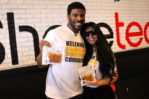 A black man and his wife, who is a woman of color wearing dark sunglasses, smiling with their arms around each other. Both are wearing white t-shirts that read kelenna's famous gourmet cornbread and holding a packaged slice of cornbread