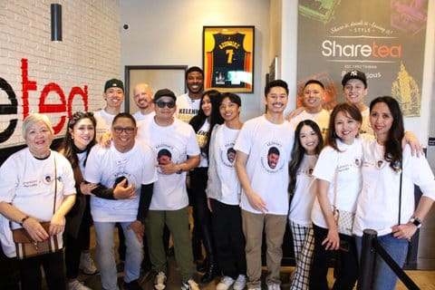 A group of several people, mostly Asian, plus one white man and one black man, all in white T-shirts, many matching. There is a framed Golden State Warriors jersey and a sign for share tea behind them
