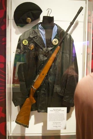 Display of a black leather jacket, black beret and shotgun behind glass. The items belonged to a member of the black panthers. There are buttons that say "Free Huey," "Free Bobby" and "cleaver for president" and one with a photo of Huey P. Newton
