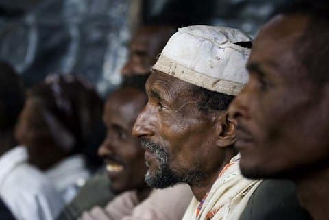 A row of Black men sitting with the focus on a man wearing a white kufi hat