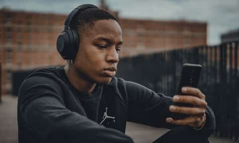 A black teen boy sitting outside with his arms resting on his knees, wearing headphones and looking at a smartphone