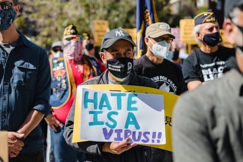 A man in a crowd wearing a gray Yankees baseball cap and black protective mask holding a sign that reads "hate is a virus" with two exclamation marks