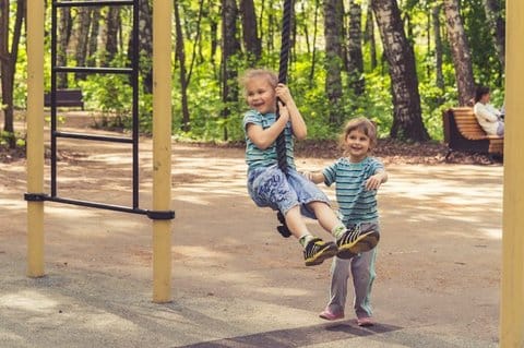 Two small children playing outdoors, with one on a rope swing and trees and a person on a bench in the background