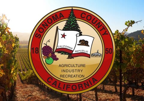 Circular logo with text that reads sonoma county california 1850 agriculture industry recreation and illustrations of a california republic flag, a green apple, a bunch of purple grapes and an evergreen tree superimposed over a photo of a vineyard