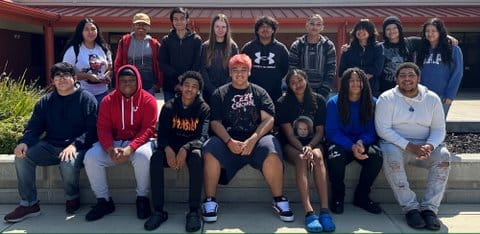 16 high school students outside their school. Seven are seated and nine are standing behind them. Most are people of color. Featured on some of their shirts are Ozzy Osbourne, Under Armour, Gap and Chucky the doll
