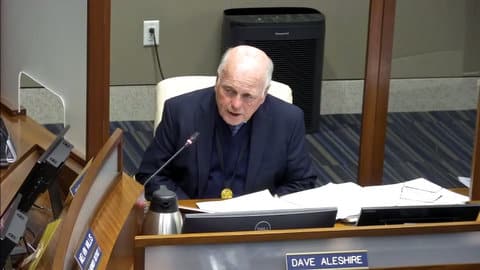 A balding white man with gray hair seated at a microphone in a government meeting. A nameplate shows he is Dave Aleshire.