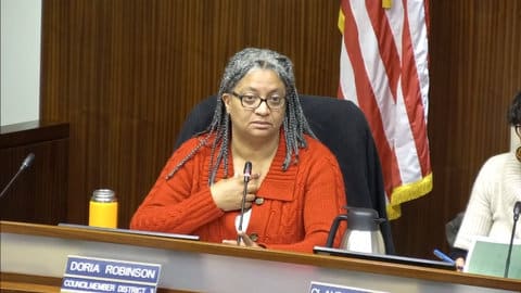 A black woman wearing glasses, red sweater and hair in braids, sitting in front of the US flag