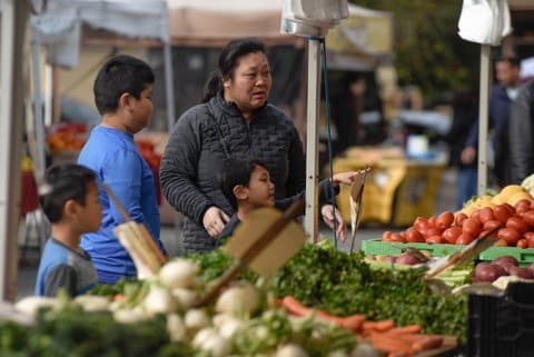 An Asian family with a woman and three boys, one of who is pointing toward the tables of fresh produce they are standing at