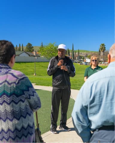A black man wearing a dark tracksuit and white hat holding a microphone in a park with green grass on a sunny day. The camera is looking at him from behind two people and a white woman is standing by him.