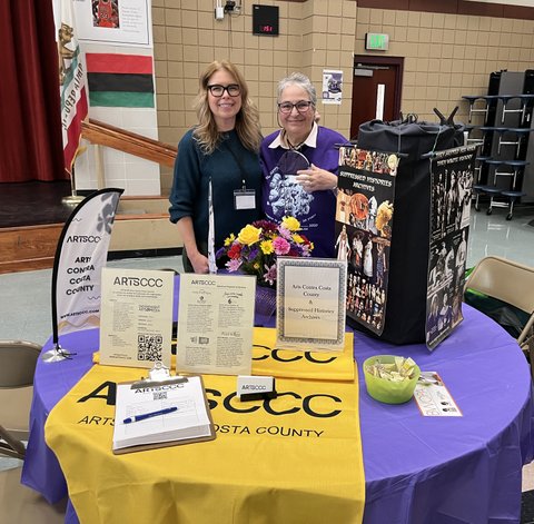 Two white women standing at a table with a purple tablecloth and yellow banner. Materials on the table says ArtsCCC and Suppressed Histories Archive.