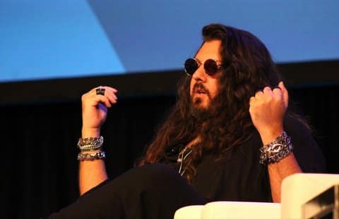 A white man with long, wavy, dark hair, goatee and soul patch, dark, round sunglasses, multiple chunky bracelets on both arms and two black rings on his right ring finger