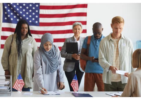 A woman wearing a headscarf is writing something on a clipboard sitting on a table with little U.S. flags on it. Also lined up at the table are a black woman, white man, black man and an Asian woman with gray hair. A large U.S. flag is on the wall behind them