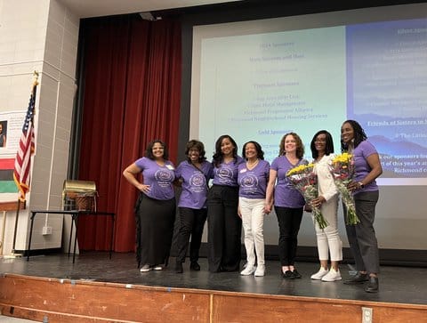 Seven women standing on a stage. Six are women of color, and six are wearing matching purple T-shirts. One black woman is dressed all in white. She and another black woman are holding bouquets of flowers