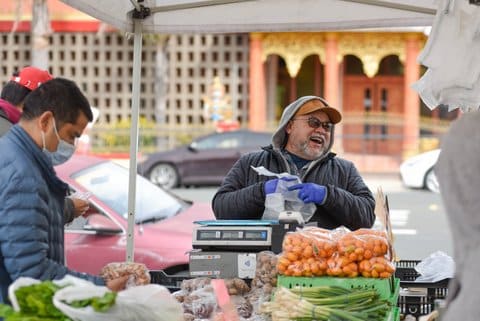 At a farmers market stand, an older Latino man wearing a jacket, hoodie, baseball cap and sunglasses laughs