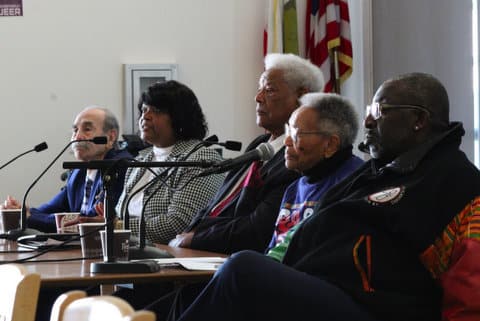An older Latino man, black woman, older black man, older black woman and black man sitting in a row at a table with microphones
