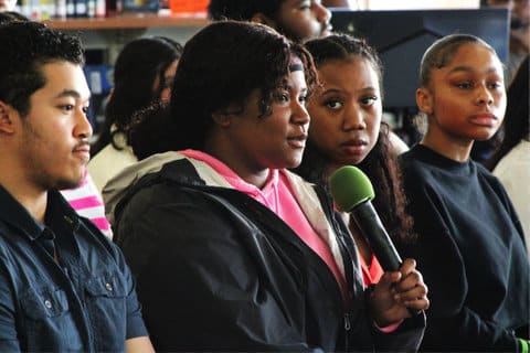 a black teen girl holding a green-topped microphone sitting among classmates