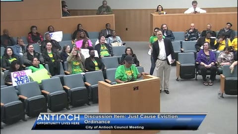 A woman in a green T-shirt speaks at a lectern at a public government meeting in front of several other people, mostly sitting and many wearing the same green shirt. On-screen text reads Antioch opportunity lives here. discussion item just cause eviction ordinance. city of antioch council meeting