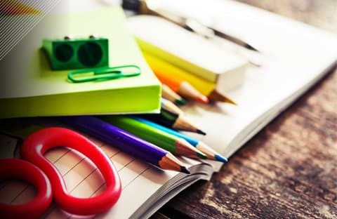 School supplies including colored pencils and a green paper clip and pencil sharpener sitting on an open notebook. The red handles of a pair of scissors are also visible