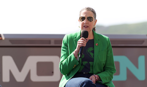 Woman wearing green jacket, black and green top and mirrored sunglasses sitting outside with microphone