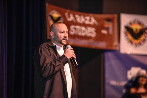 A Latino man with nearly shaved head and goatee holding microphone. An out of focus banner in the background reads la raza studies.
