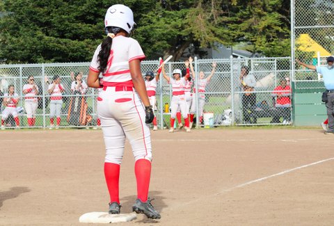 Softball player standing on third with teammates in the background cheering from the dugout