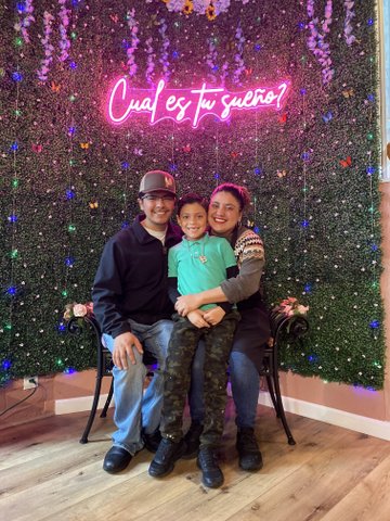 A man and woman sitting in chairs with her arm around a boy in front of them. The family is front of a wall of artificial greenery decorated with colored lights, fake butterflies and flowers and a pink neon sign that asks cual es tu sueño?